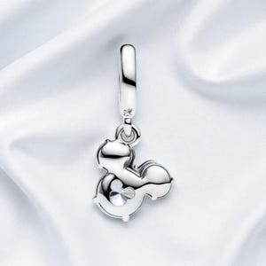 May include: A silver charm in the shape of Mickey Mouse with a clear crystal accent. The charm has a loop at the top for attaching to a bracelet or necklace. The charm is displayed on a white, silky background.