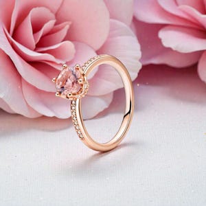 Puede incluir: Anillo de oro rosa con una gran gema rosa claro. La banda está adornada con pequeñas piedras transparentes, añadiendo brillo. El anillo se encuentra sobre un fondo floral rosa suave, creando una estética romántica y elegante. Una hermosa joya.
