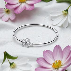 May include: A silver bangle bracelet featuring a flower-shaped charm, displayed on a white surface with pink and white flowers. The charm is adorned with small, sparkling stones, enhancing the bracelet's delicate design.