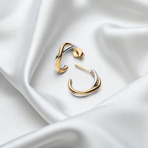 Puede incluir: Un par de pendientes de aro bicolor con un diseño entrelazado en oro y plata. Los pendientes se presentan sobre un fondo blanco y sedoso, resaltando su elegante forma y colores contrastantes.