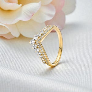 Può includere: Un anello dorato con un design a forma di V, adornato con numerose piccole pietre preziose trasparenti. L'anello è posto su uno sfondo bianco morbido con un fiore sfocato sullo sfondo. La fascia dell'anello è liscia e lucida.