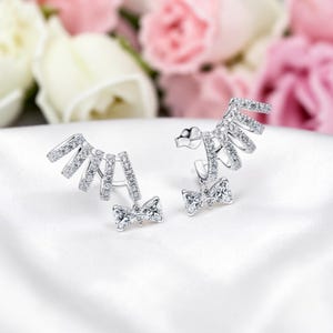 May include: A pair of silver-toned ear jackets with a bow design and multiple curved bars encrusted with small, clear gemstones. The earrings are displayed on a white surface with a blurred floral background.