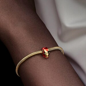 May include: A gold bracelet with a charm depicting an Iron Man helmet in red and gold. The bracelet has a snake chain design. The charm is detailed with the iconic helmet design.