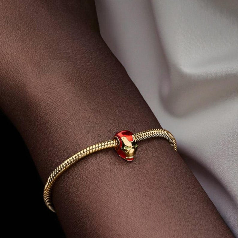 May include: A gold bracelet with a charm depicting an Iron Man helmet in red and gold. The bracelet has a snake chain design. The charm is detailed with the iconic helmet design.