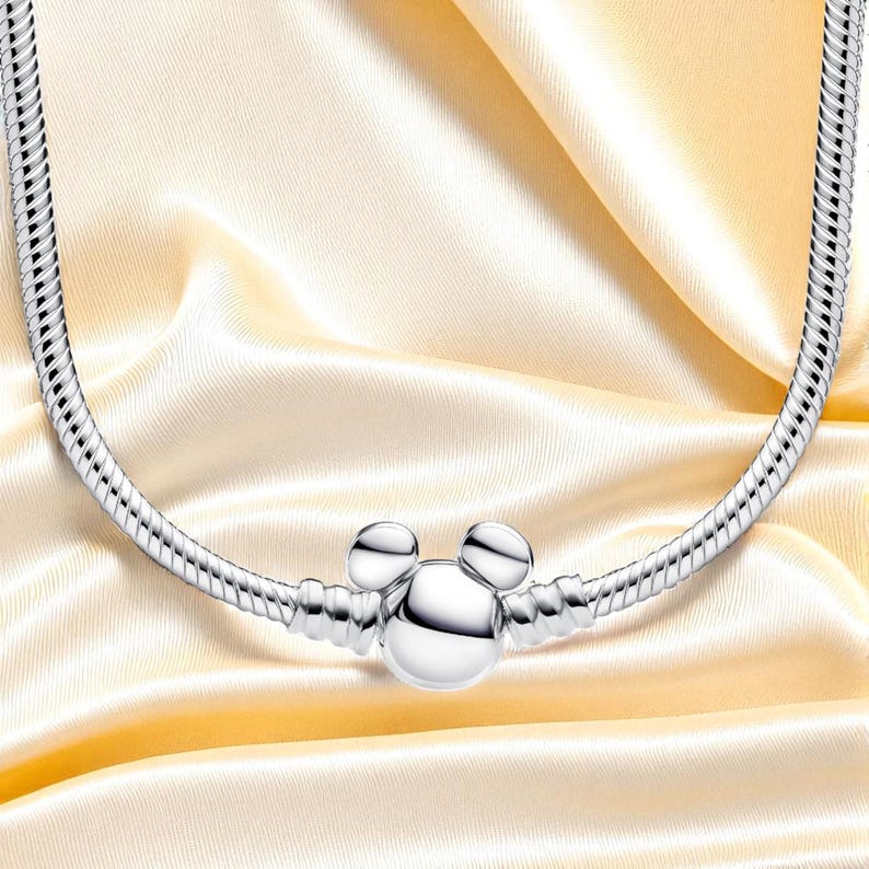 K&ouml;nnte beinhalten: Eine silberfarbene Halskette mit einem Mickey-Mouse-f&ouml;rmigen Anh&auml;nger. Die Kette hat ein Schlangenketten-Design und wird auf einem goldgelben Satin-Stoff pr&auml;sentiert. Der Anh&auml;nger hat zwei kleine Kreise f&uuml;r die Ohren und einen gr&ouml;&szlig;eren Kreis f&uuml;r das Gesicht.