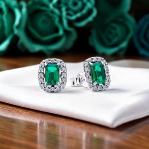 May include: A pair of emerald-cut stud earrings. Each earring has a bright green rectangular gemstone, encircled by small, clear stones. The earrings are set in silver-coloured metal and rest on a white satin surface.