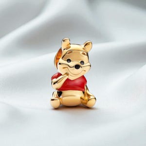 May include: A gold-colored Winnie the Pooh charm. The charm features a seated Pooh Bear wearing a red shirt. The bear has black eyes and a black nose. The charm is set against a white, silky background.