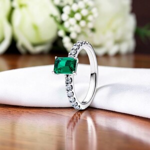 Peut inclure: Une bague en argent ornée d'une pierre verte rectangulaire taille émeraude. La monture est ornée de petites pierres précieuses claires. La bague est posée sur un tissu blanc, avec un arrière-plan floral flou.