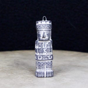 Peut inclure: Petite statue grise représentant une figure stylisée avec un corps rectangulaire et un visage détaillé. La statue a une boucle en haut pour être suspendue. Le design comprend des motifs géométriques et des figures.