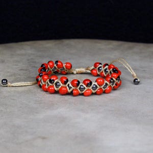 May include: A bracelet featuring two rows of small, round, bright red seeds with black spots, woven together with light tan cord. The adjustable closure includes two thin cords ending in small, dark gray metallic beads on each side.