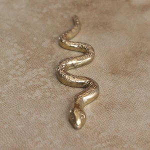 May include: A gold-toned metal snake figurine with a textured, winding body. The surface of the snake features small, repeating circular indentations along its back, creating a detailed pattern that follows the curves of its elongated, serpentine form.