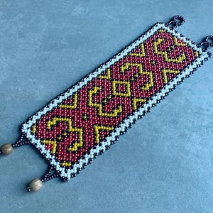 May include: A beaded bracelet with a rectangular design. The bracelet features a red background with yellow geometric patterns. The edges are outlined in white beads, and the ends have small wooden beads attached. The bracelet is approximately 5 cm wide.