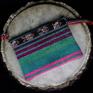 May include: A vibrant woven clutch bag with a striped pattern in shades of green, purple, and pink. The top border has a black and white geometric design with orange details. A small tassel and a woven strap are also visible.