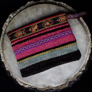 May include: A rectangular zippered pouch featuring a woven textile pattern with horizontal stripes in shades of pink, light blue, and dark brown. The top border displays a black band with a repeating orange and yellow geometric fish motif.