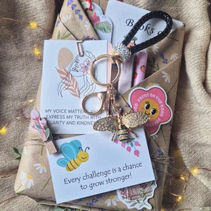 May include: A gift package wrapped in floral paper featuring a black braided keychain with a rhinestone bee charm, a card with the text 'MY VOICE MATTERS AND I EXPRESS MY TRUTH WITH CLARITY AND KINDNESS', and a card stating 'Every challenge is a chance to grow stronger!'