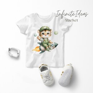 May include: White t-shirt with a cartoon tiger wearing a green hat and overalls, riding a rocket with flames. The tiger is holding a star. There are three green and white planets in the background. The text "Infinite Ideas Market" is in the top right corner.