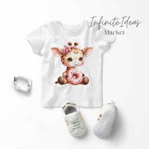 May include: White t-shirt with a cartoon giraffe wearing a pink flower crown and holding a pink donut. The giraffe is brown with a white face and black eyes. The shirt is on a white background with a pair of white shoes with gold accents.