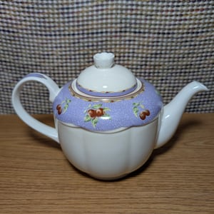 May include: A white ceramic teapot with a light purple lid and cherry blossom design. The teapot has a curved handle and spout, and a white knob on the lid. The body of the teapot is white with vertical ridges.