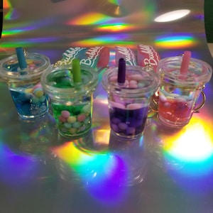 May include: Four miniature bubble tea keyrings with colourful contents and straws. The drinks are in clear plastic cups with gold-coloured handles. The background is a reflective surface with rainbow-coloured light.