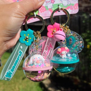 May include: Two pink and green glitter filled keychain ornaments featuring Sanrio characters. The keychain is attached to a green strap with the word "Cinnamoroll" printed on it. The pink ornament features Hello Kitty and the green ornament features My Melody.