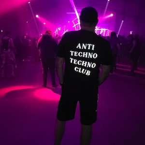 May include: A person wearing a black t-shirt with the text "ANTI TECHNO TECHNO CLUB" in white letters. The person is standing in a dark room with purple and pink lights.