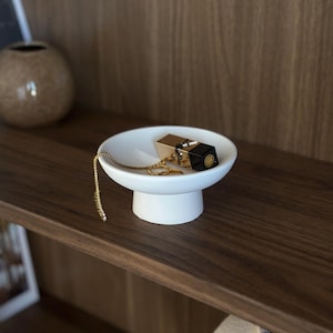 Peut inclure: Un bol sur pied en céramique blanche contenant un tube de rouge à lèvres doré et un collier à chaîne dorée délicat. Le bol présente un bassin large et peu profond soutenu par une base cylindrique robuste, offrant un support simple et épuré.