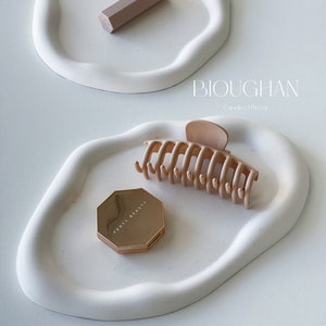Peut inclure: Deux plateaux en céramique blanche aux bords irréguliers, l'un sur l'autre. Le plateau supérieur contient un compact de maquillage Fenty Beauty doré, une pince à cheveux marron clair et le texte "BIOUGHAN Candles | Decor".