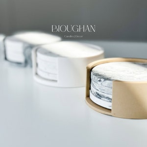 Peut inclure: Ensemble de dessous de verre à motif marbré dans un support blanc et beige. Les dessous de verre présentent un motif tourbillonnant gris et blanc. Le support est en forme de demi-cercle. Le texte "BIOUGHAN Candles | Decor" est visible.