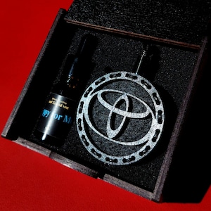 Könnte beinhalten: Eine dunkle Holzbox enthält einen silberfarbenen Toyota-Logo-Anhänger und eine schwarze Flasche. Auf der Flasche steht in blauer Schrift "Happy for M". Der Anhänger ist rund mit einem dekorativen Rand.