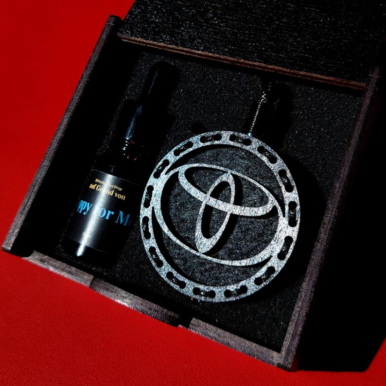 Puede incluir: Una caja de madera oscura contiene un colgante plateado con el logotipo de Toyota y una botella negra. La botella tiene texto azul que dice "Happy for M". El colgante es redondo con un borde decorativo.