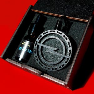 Könnte beinhalten: Eine schwarze Holzkiste mit dunkelgrauem Innenraum enthält einen schwarzen Anhänger mit Blitzdesign und eine kleine Glasflasche. Die Flasche hat einen schwarzen Deckel und ein Etikett mit der Aufschrift "Happy for Me."