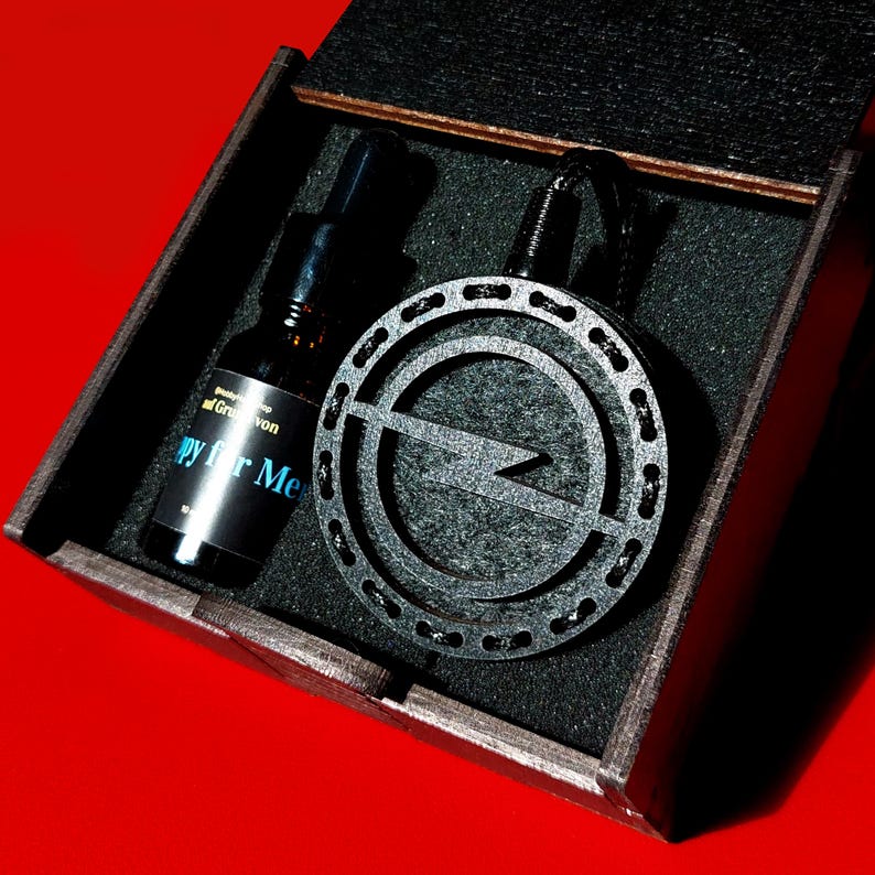 May include: A black wooden box with a dark grey interior contains a black pendant with a lightning bolt design and a small glass bottle. The bottle has a black cap and a label that reads "Happy for Me."