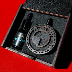 Könnte beinhalten: Eine dunkle Holzbox enthält einen BMW-Logo-Anhänger und eine kleine Flasche. Der Anhänger ist ein kreisförmiges Design mit dem BMW-Emblem. Die Flasche hat ein schwarzes Etikett mit dem Text "Happy for Me".