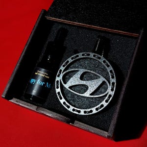 Könnte beinhalten: Eine schwarze Holzbox enthält einen silberfarbenen Anhänger mit dem Hyundai-Logo und eine schwarze Flasche mit dem Text "Happy for Me". Die Box steht auf einer roten Oberfläche.