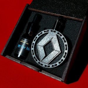 Könnte beinhalten: Eine schwarze Holzkiste mit einem silbernen und schwarzen geometrischen Design-Anhänger und einer kleinen Flasche mit einem schwarzen Etikett, auf dem "Grand von" und "Happy for Me" steht.