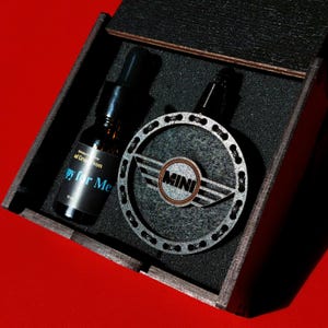 Könnte beinhalten: Eine dunkle Holzbox enthält eine schwarze Flasche mit dem Text "py for Me" und ein silbernes Mini-Emblem. Das Emblem ist ein kreisförmiges Design mit dem Wort "MINI" in der Mitte. Der Hintergrund ist rot.
