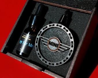 Könnte beinhalten: Eine dunkle Holzbox enthält eine schwarze Flasche mit dem Text "py for Me" und ein silbernes Mini-Emblem. Das Emblem ist ein kreisförmiges Design mit dem Wort "MINI" in der Mitte. Der Hintergrund ist rot.