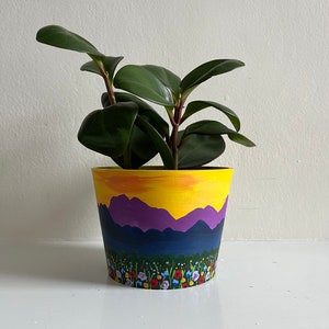 May include: A yellow, purple, and blue painted ceramic planter with a sunset scene and a field of flowers. A green leafy plant is growing in the planter.