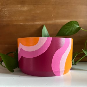 May include: A hand-painted ceramic planter with a retro design. The planter features a curved pattern in shades of orange, pink, and burgundy. The planter is a small, round shape, perfect for small plants. The background is a wooden surface.