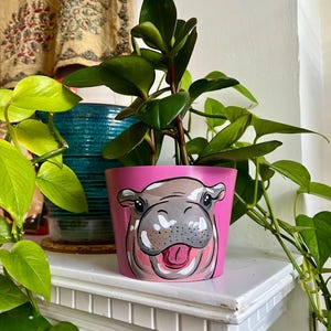 May include: A pink painted ceramic flower pot featuring a smiling hippo face. The pot holds a green leafy plant. The pot is sitting on a white shelf, with other plants and a patterned curtain in the background.