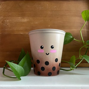 May include: A white plastic cup with a brown ombre design and black polka dots. The cup has a painted face with pink cheeks and a smile.