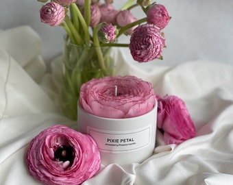 Pode incluir: Uma vela rosa Pixie Petal com aroma de morango, frésia e baunilha, em um recipiente branco. A vela é cercada por flores rosas e um vaso de flores rosas. A vela tem um pavio.