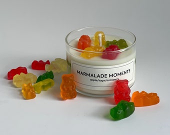 Pode incluir: Uma vela branca em um pote de vidro transparente com um rótulo que diz "Marmalade Moments apple/sugar/cranberry". A vela é coberta por ursinhos de goma de várias cores, incluindo vermelho, laranja, amarelo e verde.