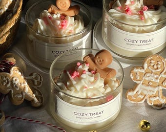 May include: Three glass jars with white candle wax and a gingerbread man figurine on top. The jars have labels that say "Cozy Treat" and "ginger/sugar/butter".