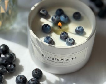 Pode incluir: Uma vela de cerâmica branca com o texto "BLUEBERRY BLISS" e mirtilos frescos espalhados por cima. A vela está acesa e a chama é visível. A vela é rodeada por mais mirtilos.