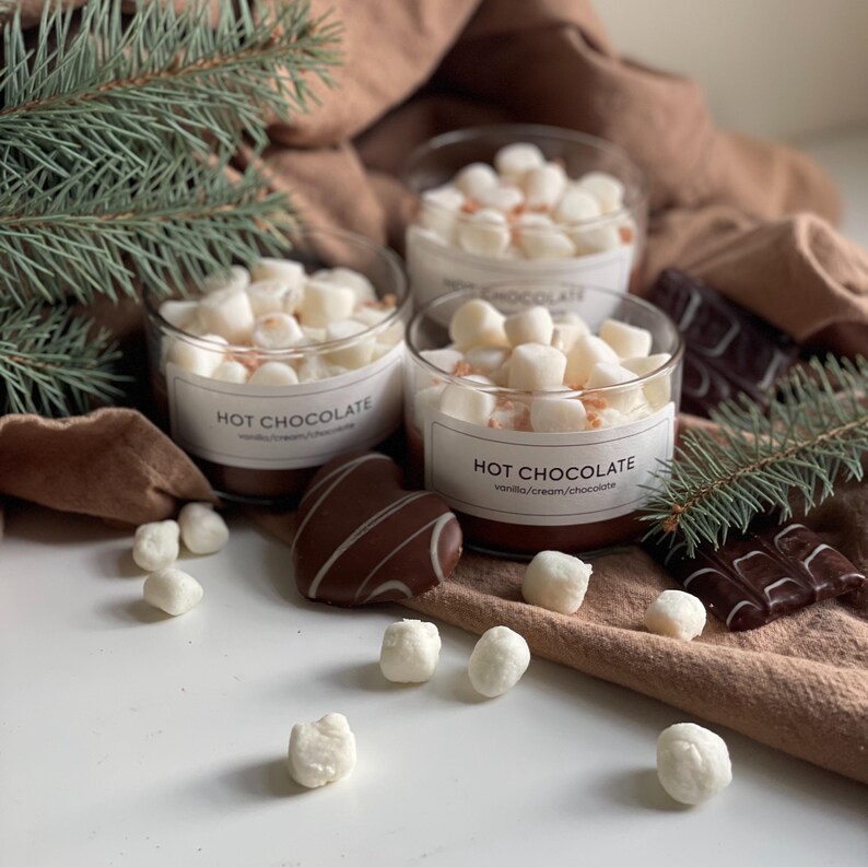 Pode incluir: Tr&ecirc;s frascos de vidro cheios de chocolate quente, cobertos com marshmallows. Cada frasco tem um r&oacute;tulo que diz "HOT CHOCOLATE" com "vanilla/cream/chocolate" abaixo. Bombons de chocolate e um ramo de pinheiro tamb&eacute;m est&atilde;o presentes.