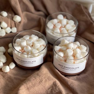 Pode incluir: Tr&ecirc;s velas de vidro transparente preenchidas com cera branca, cobertas com marshmallows e granulado. Cada vela tem um r&oacute;tulo que diz "HOT CHOCOLATE" com "vanilla/cream/chocolate" abaixo. Sobre um tecido marrom.