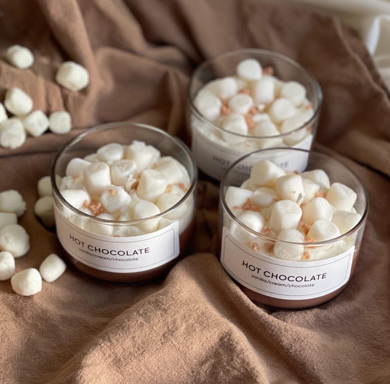 Pode incluir: Tr&ecirc;s velas de vidro transparente preenchidas com cera branca, cobertas com marshmallows e granulado. Cada vela tem um r&oacute;tulo que diz "HOT CHOCOLATE" com "vanilla/cream/chocolate" abaixo. Sobre um tecido marrom.