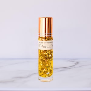 May include: A clear glass bottle with a gold cap filled with a yellow liquid and dried flowers. The bottle has a white label with the text "mystic lunamagic focus".