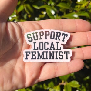 Puede incluir: Una pegatina rectangular con el texto "SUPPORT LOCAL FEMINIST" en negro, contorneado en blanco, sobre un fondo rosa. La pegatina se sostiene en una mano sobre un fondo de follaje verde.